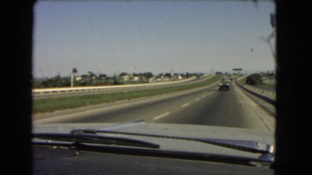 1966 California windshield POV of a car traveling on a broad highway beside a grassy median and low roadside housing.