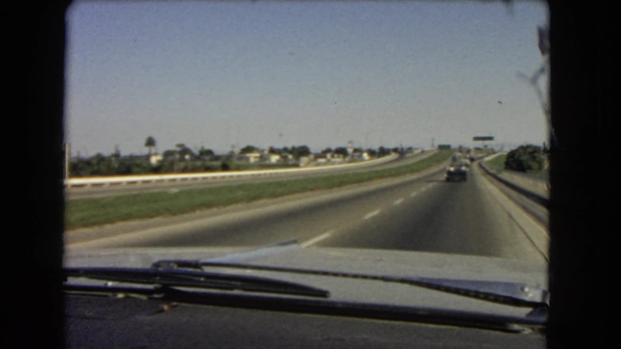 1966 California 8mm frame from inside a moving car, showing highway lanes, a grassy center strip, distant housing, and dashboard detail.