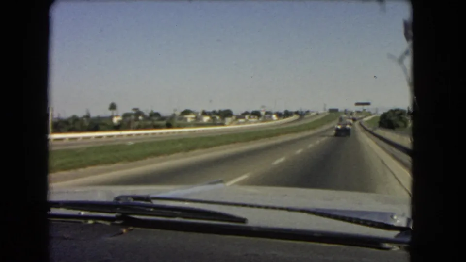 1966 California 8mm frame from inside a moving car, showing highway lanes, a grassy center strip, distant housing, and dashboard detail.