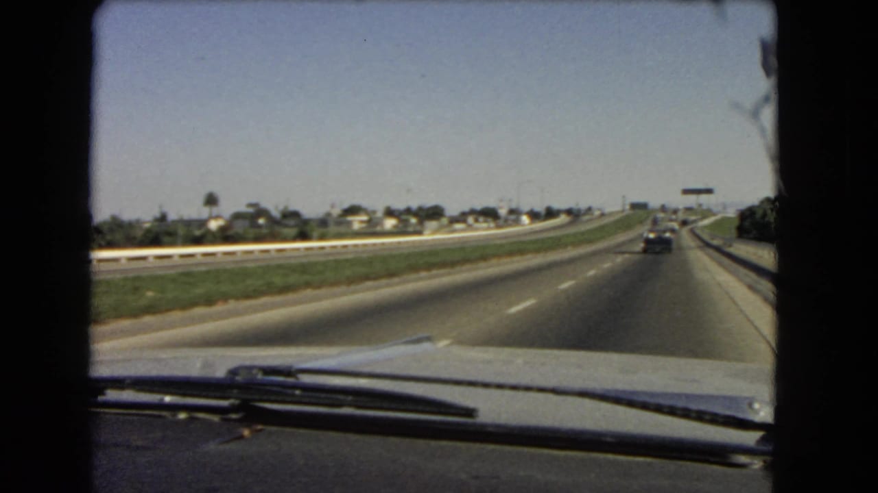 1966 California home-movie still of highway driving, with the windshield wipers, dashboard, grass median, and distant roadside buildings visible.
