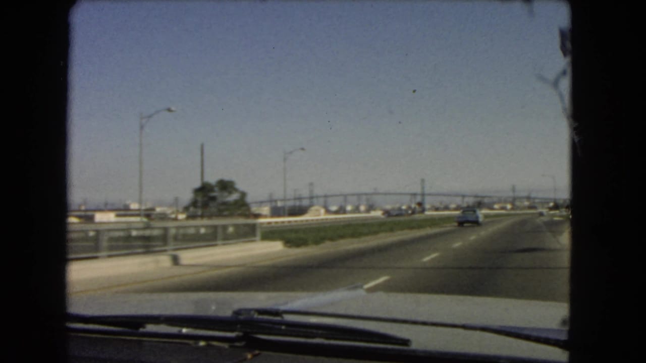 Passenger-side windshield view on a 1966 California highway, with a car ahead, roadside barriers, streetlights, and distant bridge-like spans.