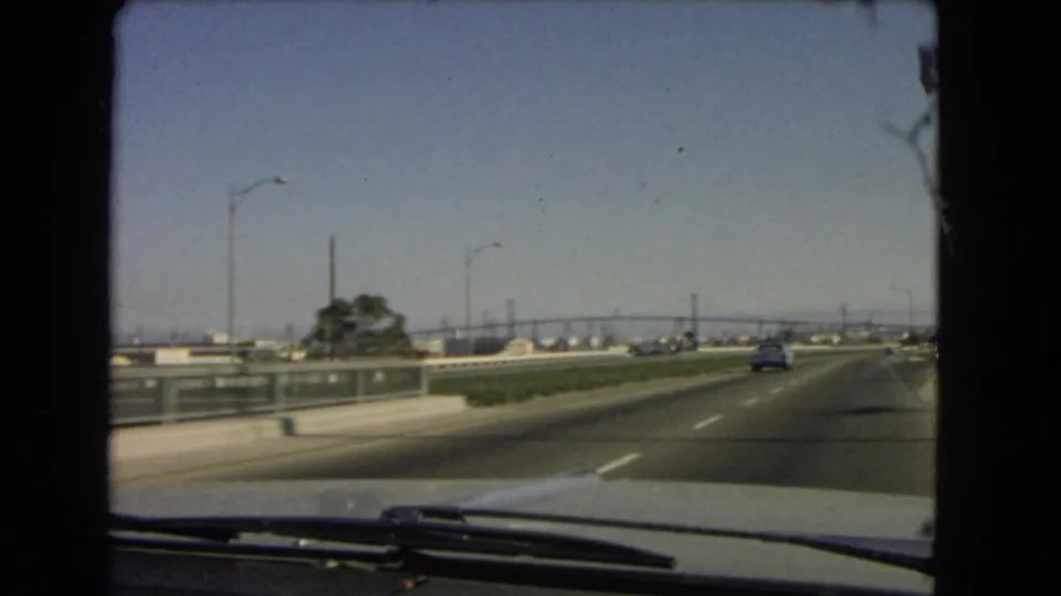 Passenger-side windshield view on a 1966 California highway, with a car ahead, roadside barriers, streetlights, and distant bridge-like spans.
