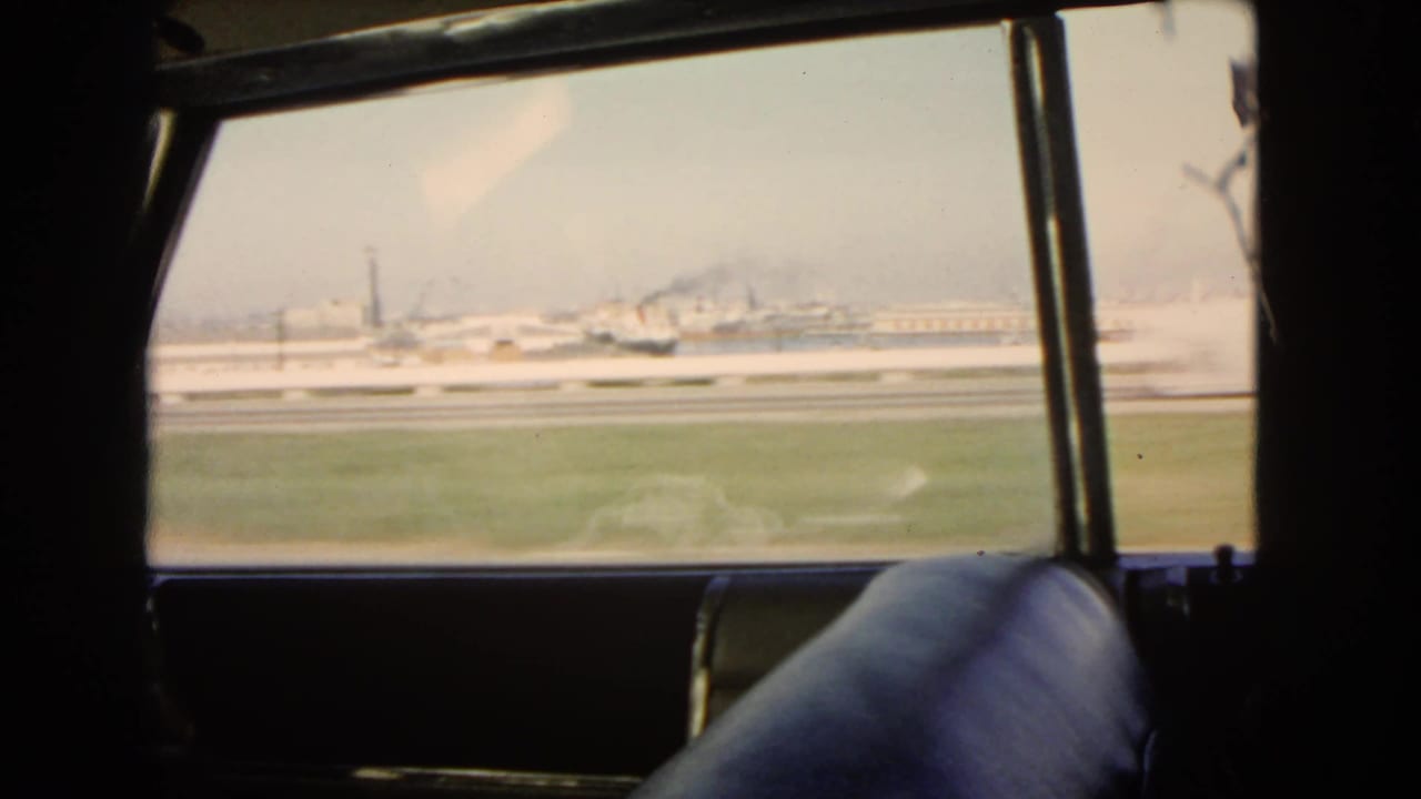 1966 California side-window travel view, with the car interior in silhouette, grassy verge, roadway barriers, and distant industrial buildings or stacks.