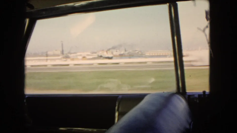 1966 California side-window travel view, with the car interior in silhouette, grassy verge, roadway barriers, and distant industrial buildings or stacks.