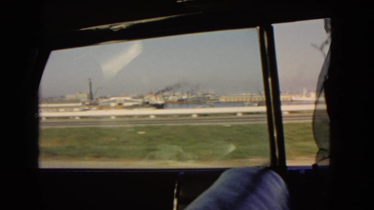 Moving side-window view from a 1966 California car trip, showing a grassy roadside, roadway barriers, industrial structures, and part of the passenger seat.