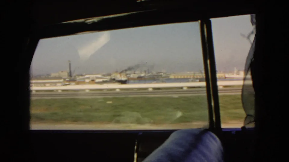 Moving side-window view from a 1966 California car trip, showing a grassy roadside, roadway barriers, industrial structures, and part of the passenger seat.