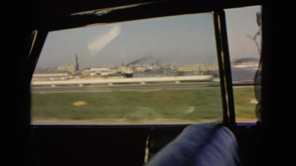 1966 California car-window view over roadside grass toward a harbor, distant buildings, water, and a smokestack.