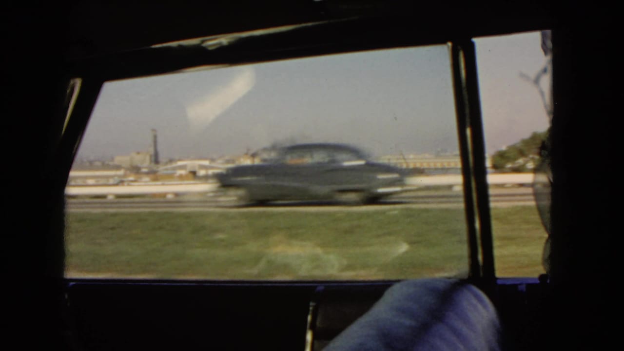 A dark mid-century sedan passes the window during a 1966 California harbor-side drive, with water and buildings beyond.