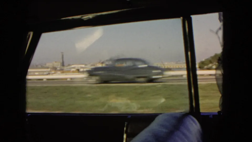 A dark mid-century sedan passes the window during a 1966 California harbor-side drive, with water and buildings beyond.
