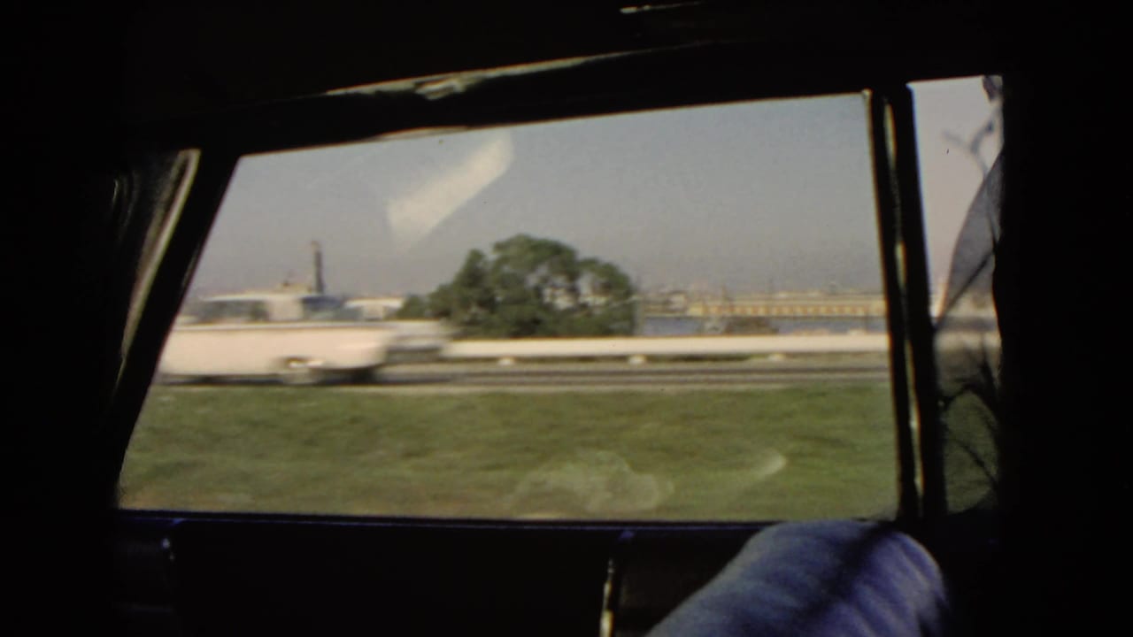 A white car blurs past the passenger window on a 1966 California coastal road, with a tree and harbor buildings behind.