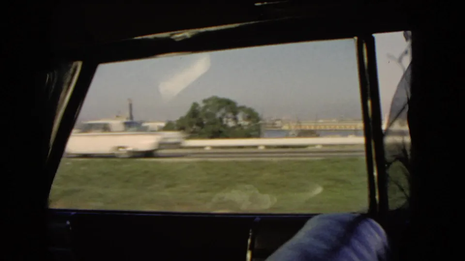 A white car blurs past the passenger window on a 1966 California coastal road, with a tree and harbor buildings behind.