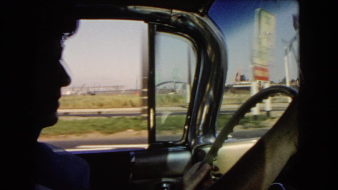 Interior car view in California, 1966, with the driver in profile and roadside signs and industrial shapes beyond the window.
