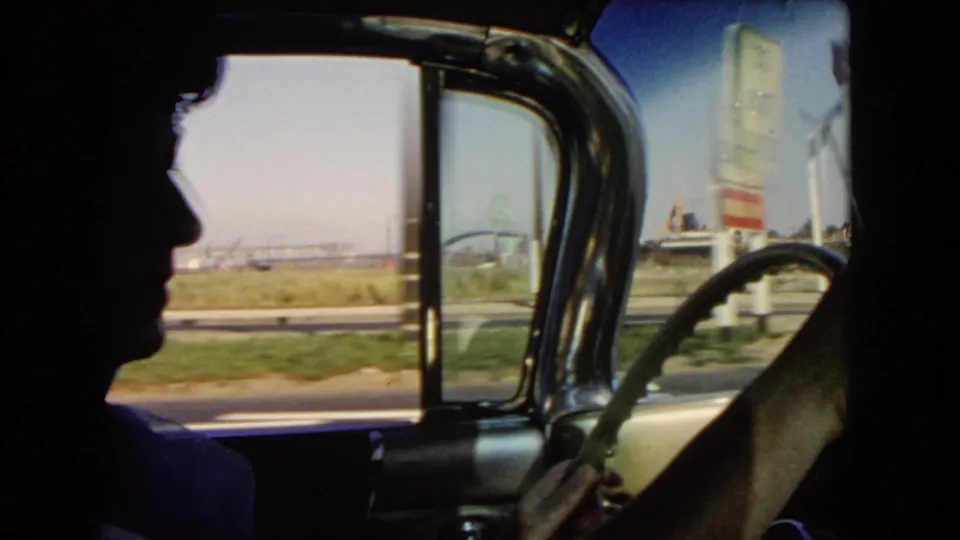 Interior car view in California, 1966, with the driver in profile and roadside signs and industrial shapes beyond the window.