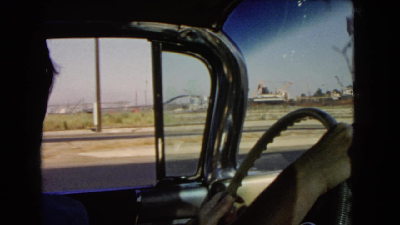 1966 California home-movie frame looking past the driver’s hands and steering wheel toward roadside poles and distant industrial structures.