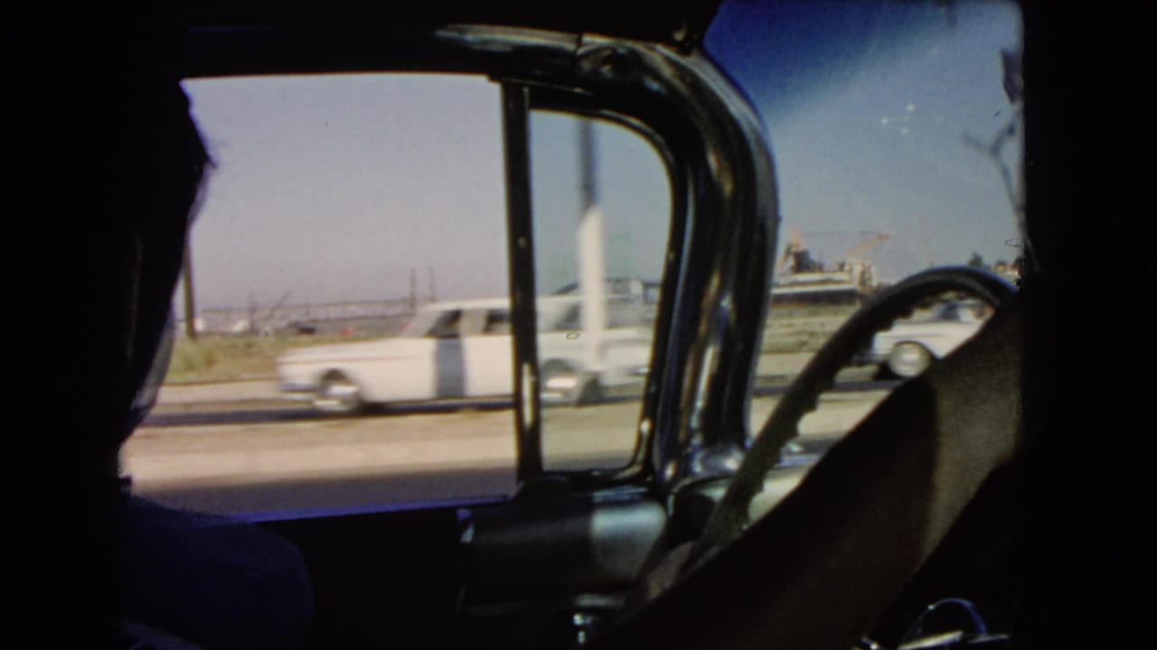 1966 California 8mm frame from inside a classic car, with a driver's silhouette and passing highway traffic beyond the window.