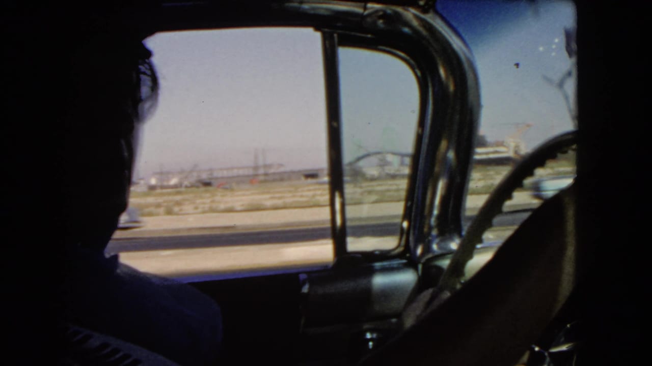 Interior 1960s car view in California, showing the driver in silhouette against a bright highway and distant industrial structures.