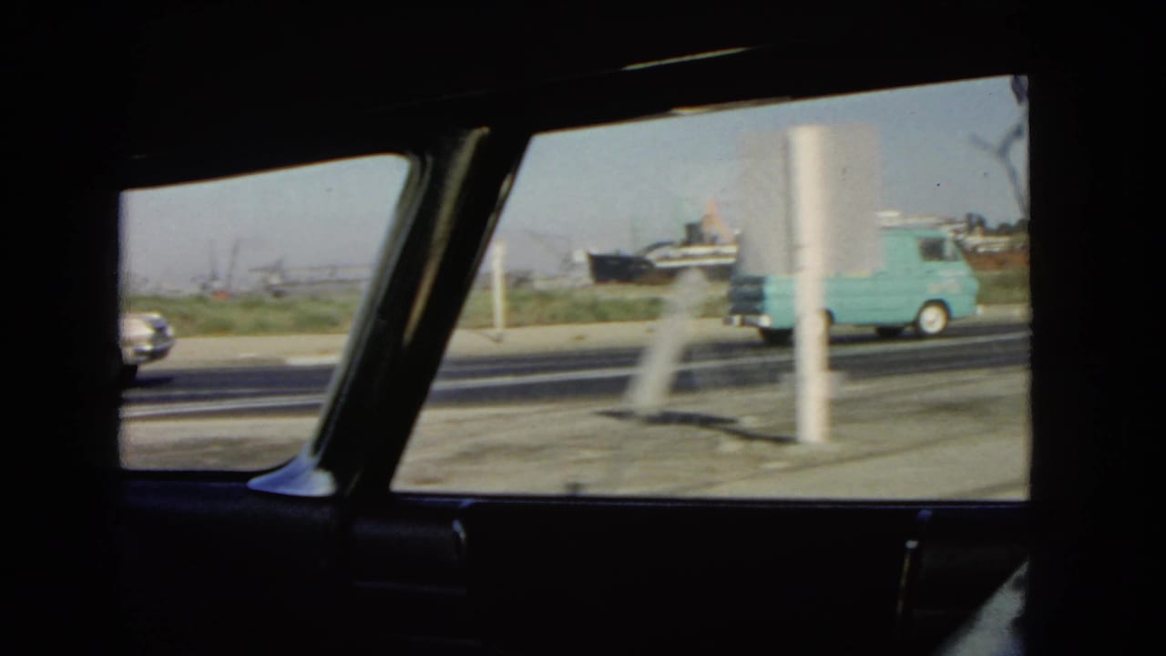 1966 California road traffic viewed from inside a moving car, with a pale blue delivery van, sedans, roadside poles, and distant industrial structures.