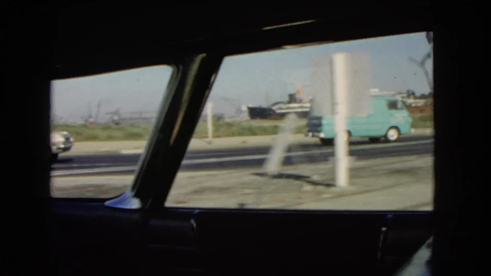 1966 California road traffic viewed from inside a moving car, with a pale blue delivery van, sedans, roadside poles, and distant industrial structures.