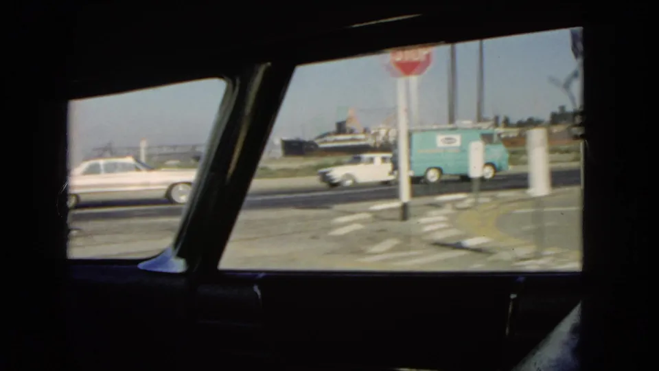 Roadside traffic in California in 1966 seen through a car window, with a white sedan, light blue delivery van, stop sign, and distant ships or industrial equipment.