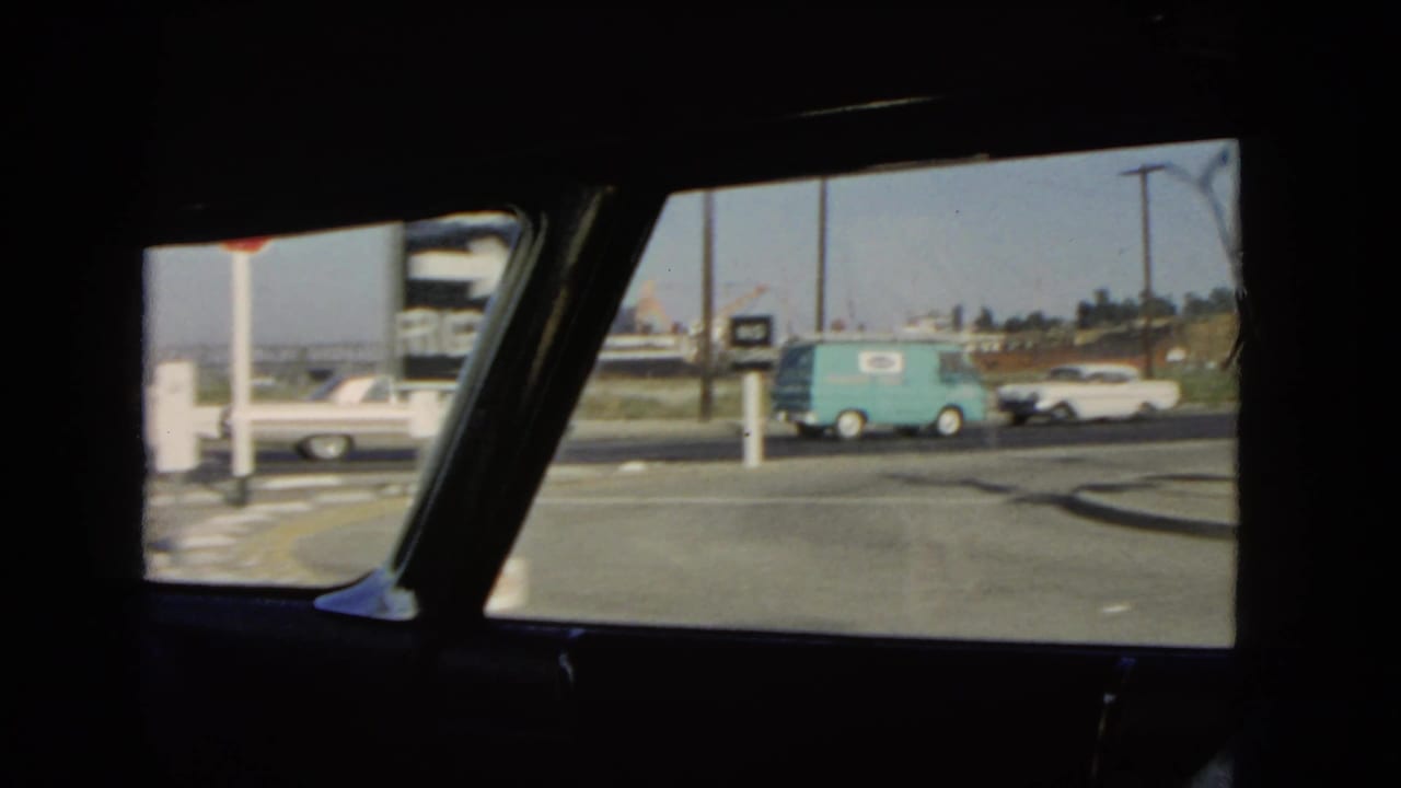 Passenger-side view of a 1966 California street with a blue delivery van, white sedan, road signs, curb, utility poles, and industrial background details.