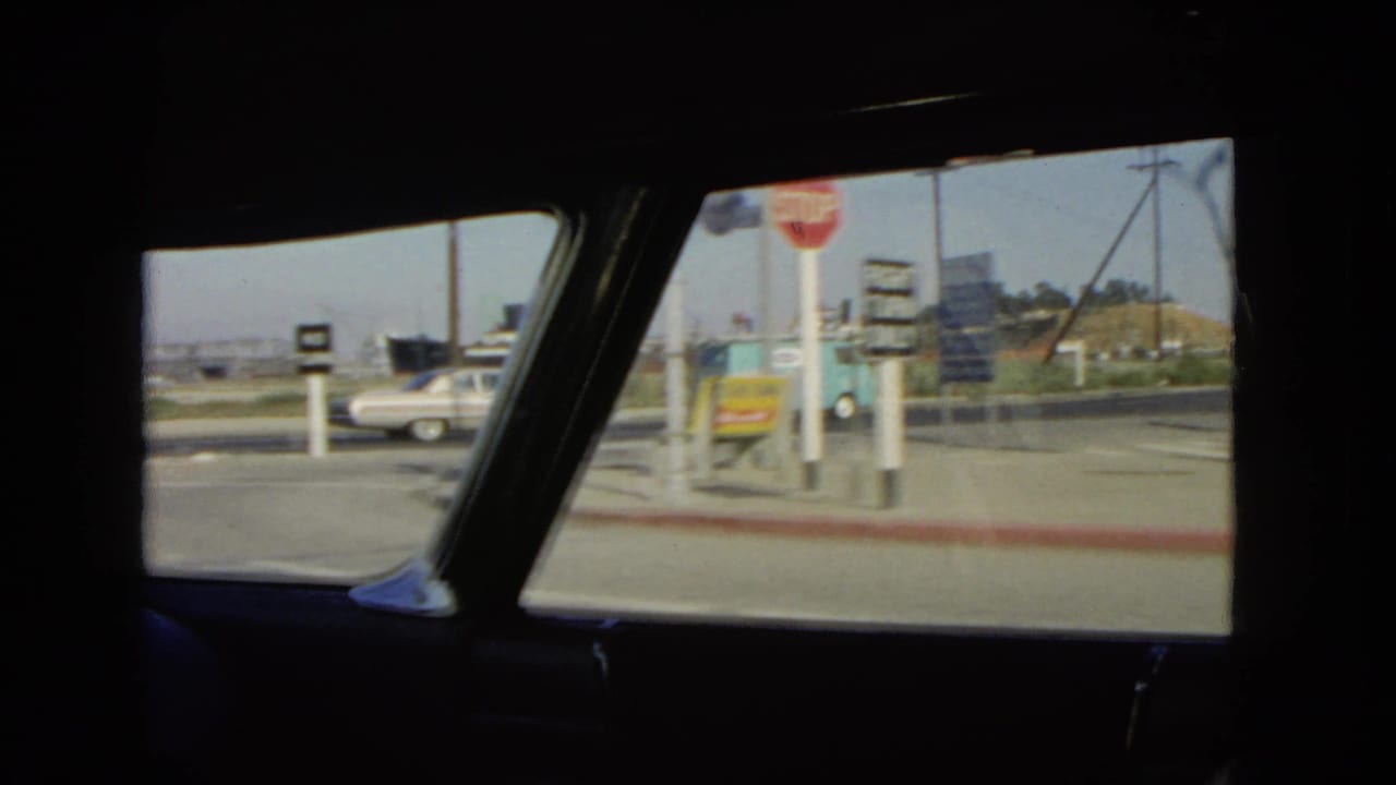 1966 California roadside scene from inside a moving car, showing a red stop sign, signboard, white sedan, blue van, curb, and industrial waterfront background.