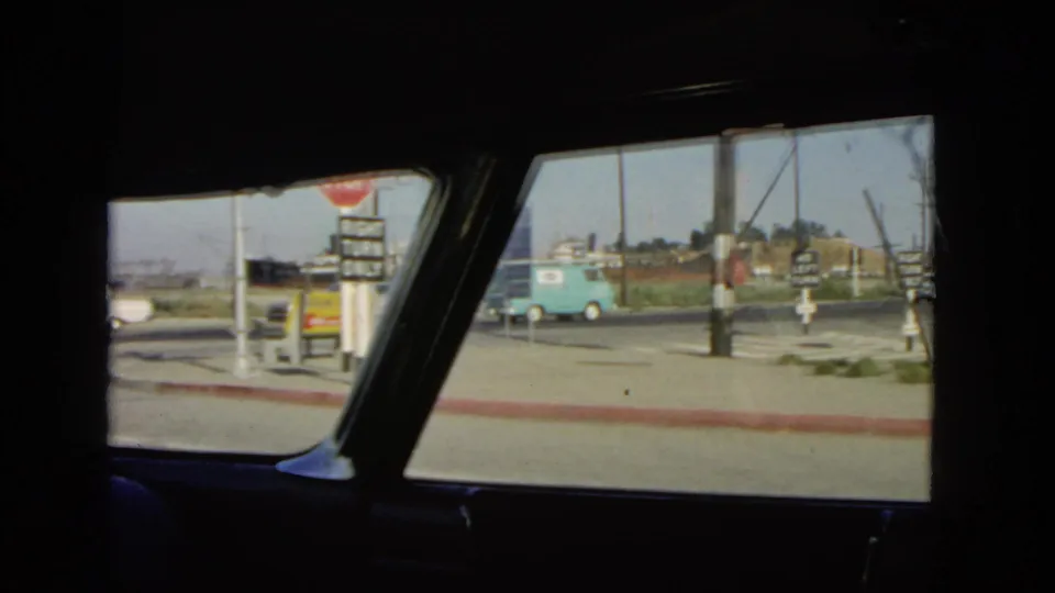 1966 California home-movie view through a car window toward roadside signs, traffic, and a turquoise van.