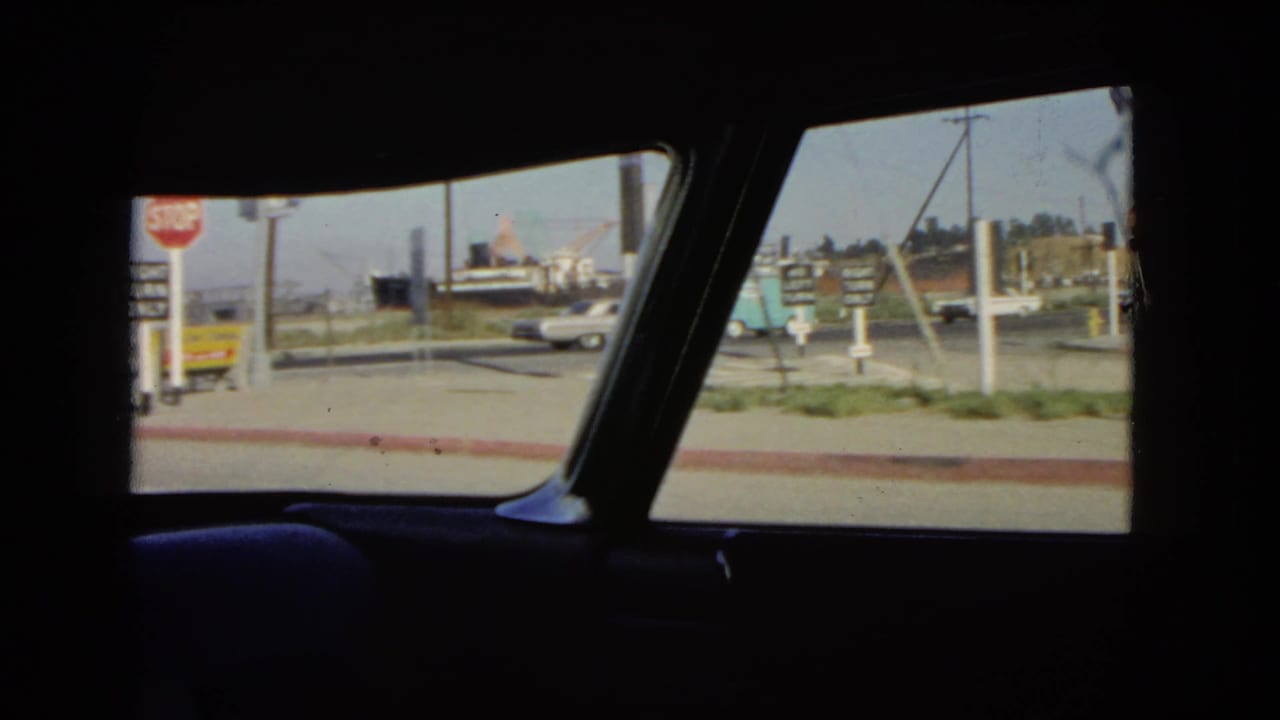 1966 California street scene filmed from inside a car, with roadside signs, a white sedan, and low buildings.