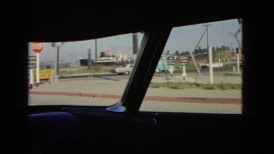 1966 California street scene filmed from inside a car, with roadside signs, a white sedan, and low buildings.