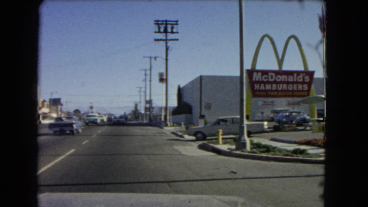 1966 California home-movie frame of traffic passing a McDonald's golden arches sign and roadside parking.