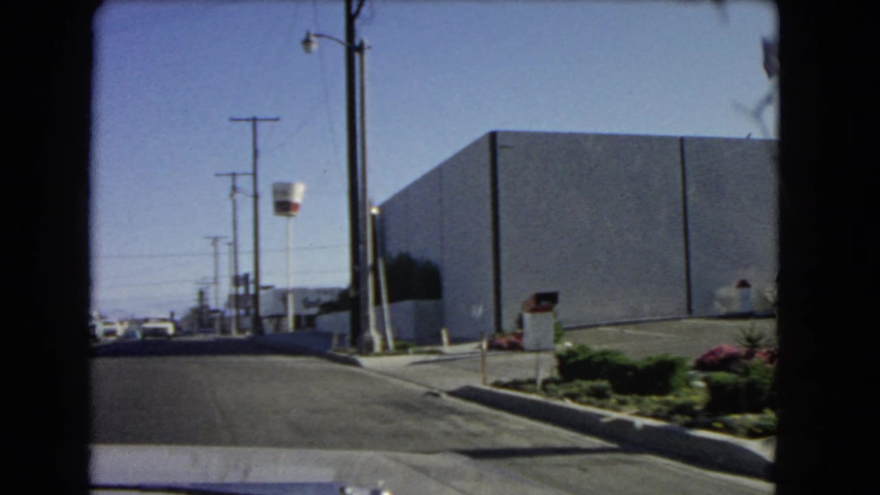 1966 California car-window view along a commercial street with a large building, utility poles, curbside planting, and distant vehicles.