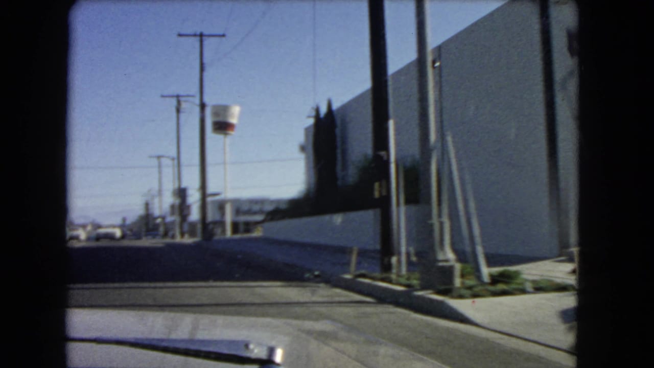 1966 California car-window view passing a white wall, sidewalk, utility poles, and low roadside buildings under a clear sky.