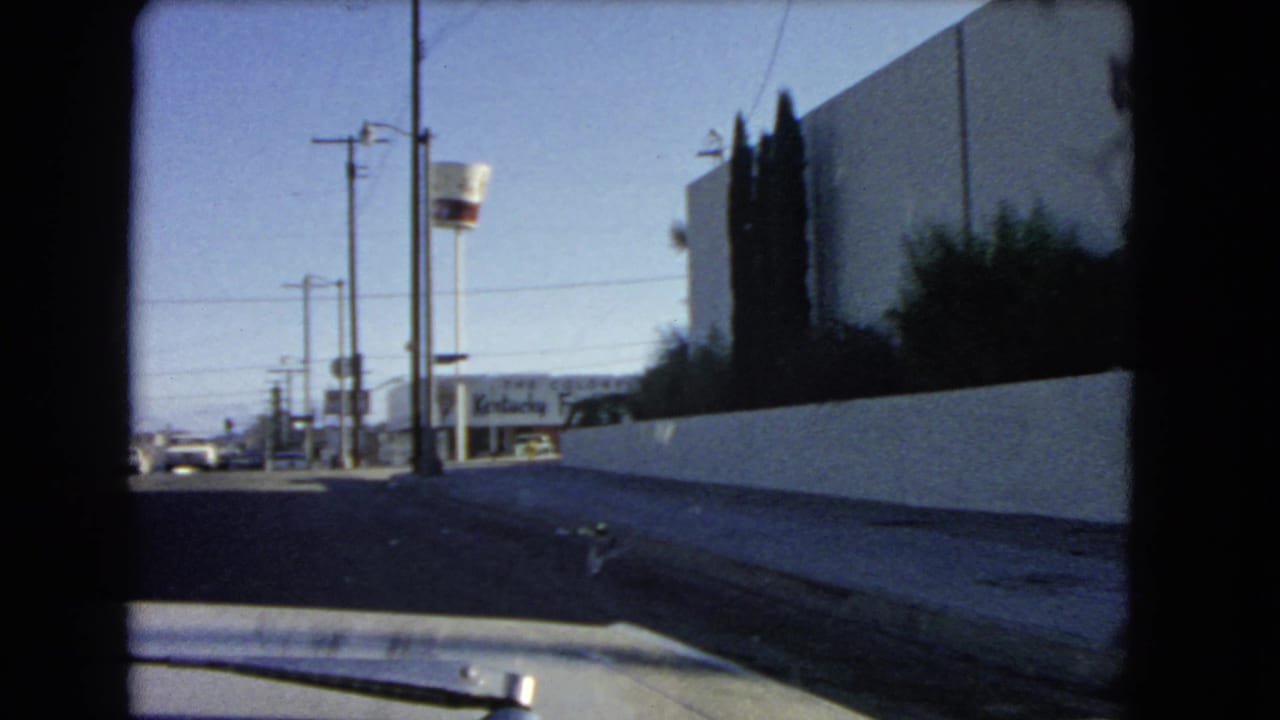 Moving-car view along a white wall toward roadside signs and low commercial buildings.