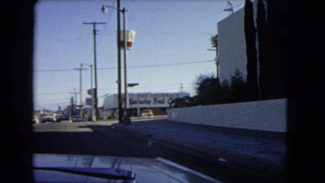 1966 California drive-by frame showing utility poles, a white wall, sidewalk, cars, and a distant Kentucky Fried Chicken sign.