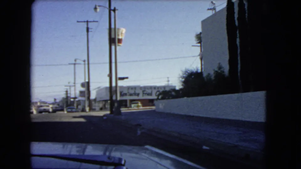 1966 California drive-by frame showing utility poles, a white wall, sidewalk, cars, and a distant Kentucky Fried Chicken sign.