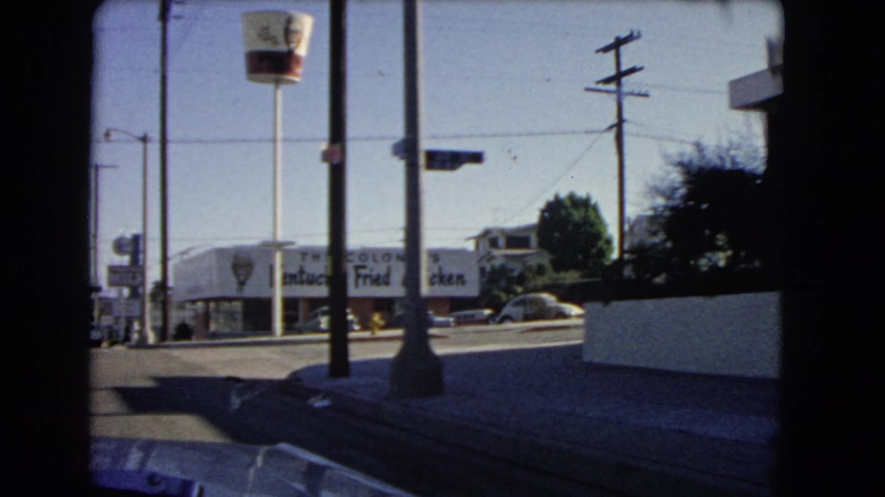 California street seen from a car with KFC signage, utility poles, sidewalk, and mid-century cars.