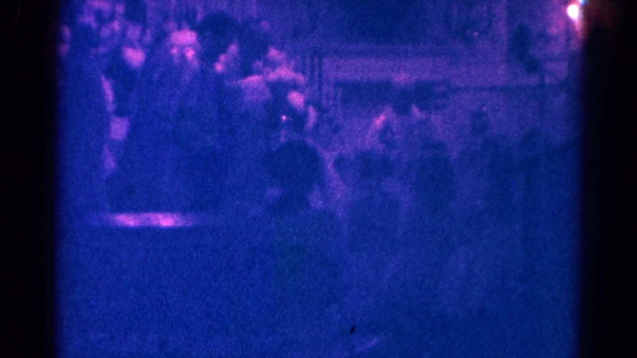 Low-light 1966 Danville home-movie frame with several people seated in a crowded interior and a bright light near the right edge.