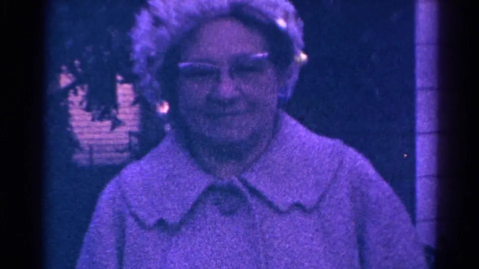 1966 Danville, Illinois home-movie frame of an adult woman in glasses and a light collared garment near house siding.