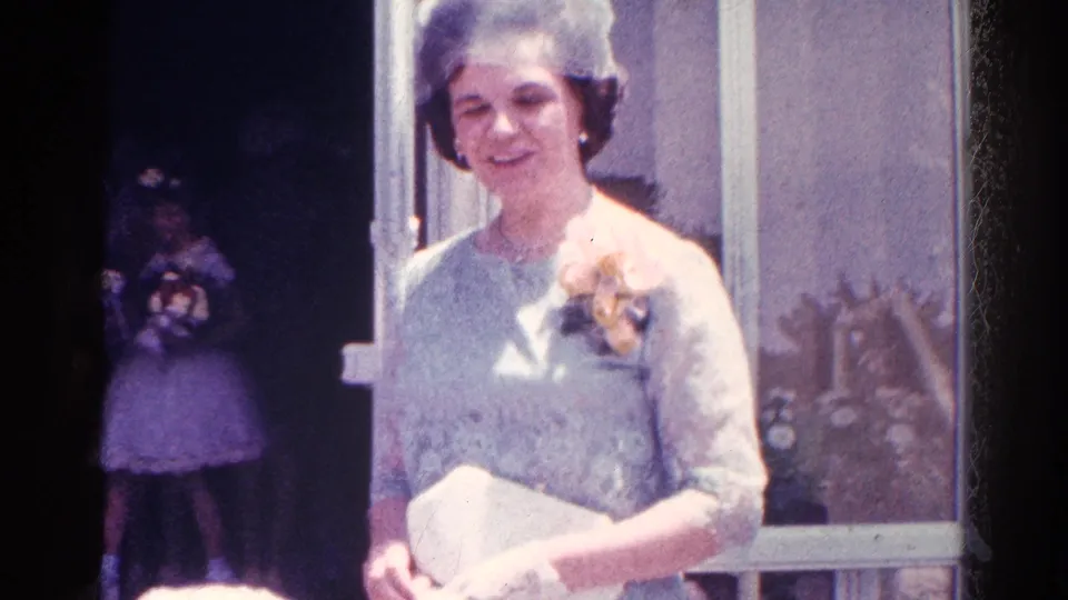 A formally dressed woman with hat, corsage, and clutch stands near the doorway while another guest appears in the shaded entrance.