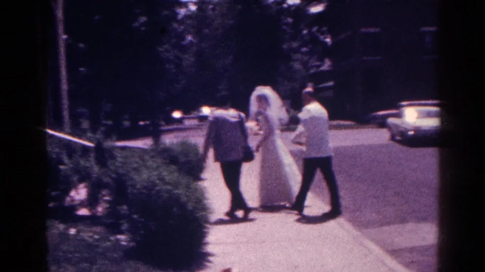 Bride in a white gown walks along a sidewalk with two adults in 1966 Danville, Illinois home-movie footage.