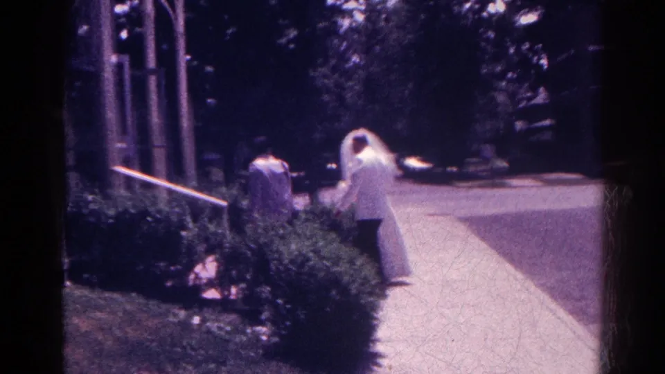 The veiled bride and companions continue down a sidewalk near hedges and a street in Danville wedding footage.
