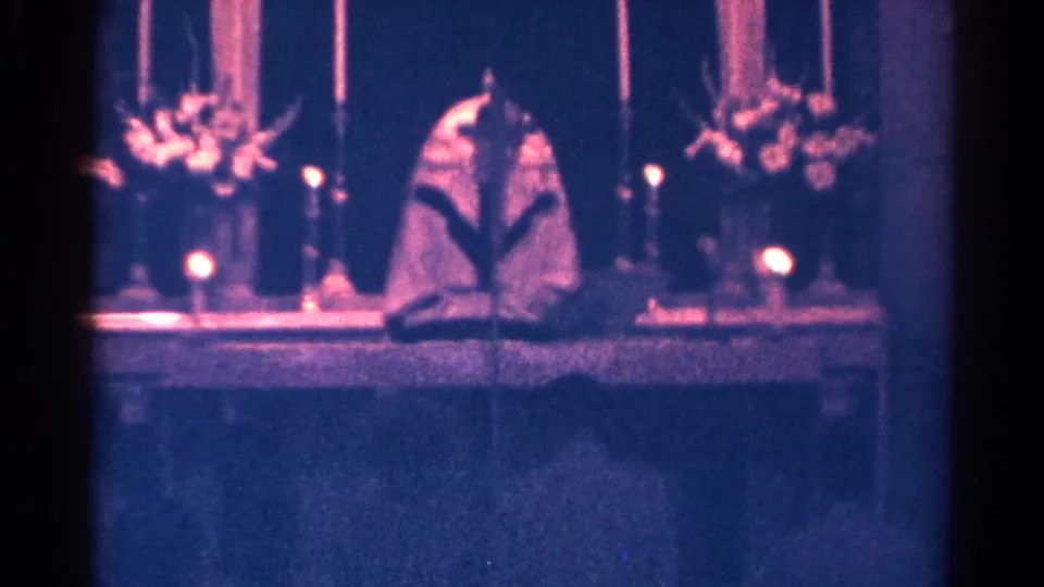 The 8mm frame shows the pastor reading at the altar, with candles, flowers, and dark vertical architectural details behind him.