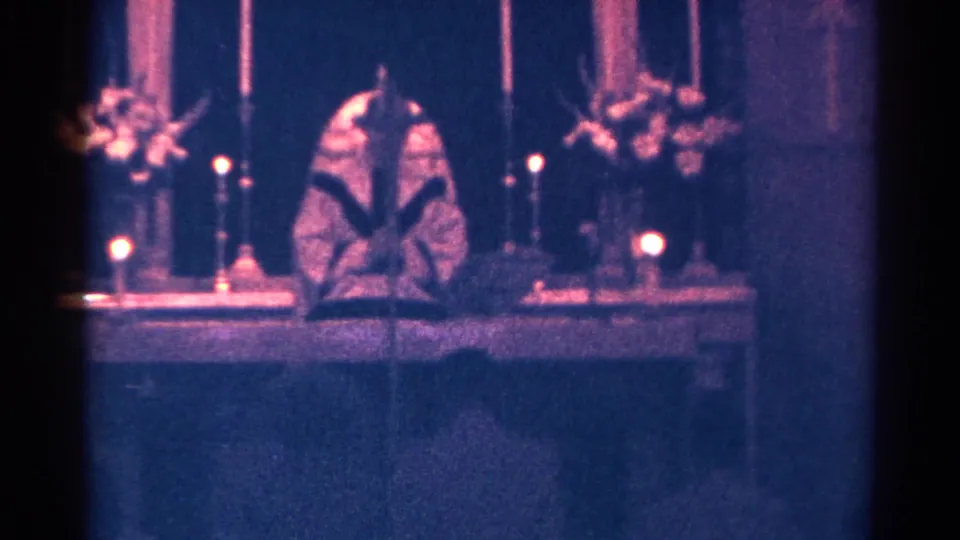 A static 1966 home-movie frame holds on the pastor at the candlelit altar, with flowers and an open book visible.