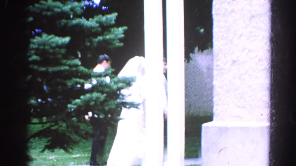 In the 1966 Danville wedding footage, a bride in white appears beside men in white suits near columns, grass, and an evergreen tree.