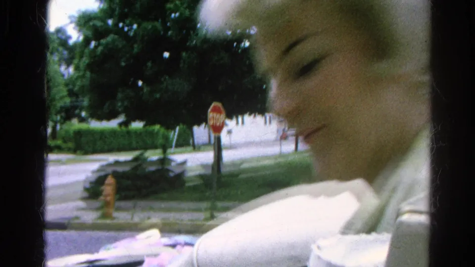 A motion-blurred close view of a blonde woman passes the camera, with a stop sign, sidewalk, and residential street behind her.