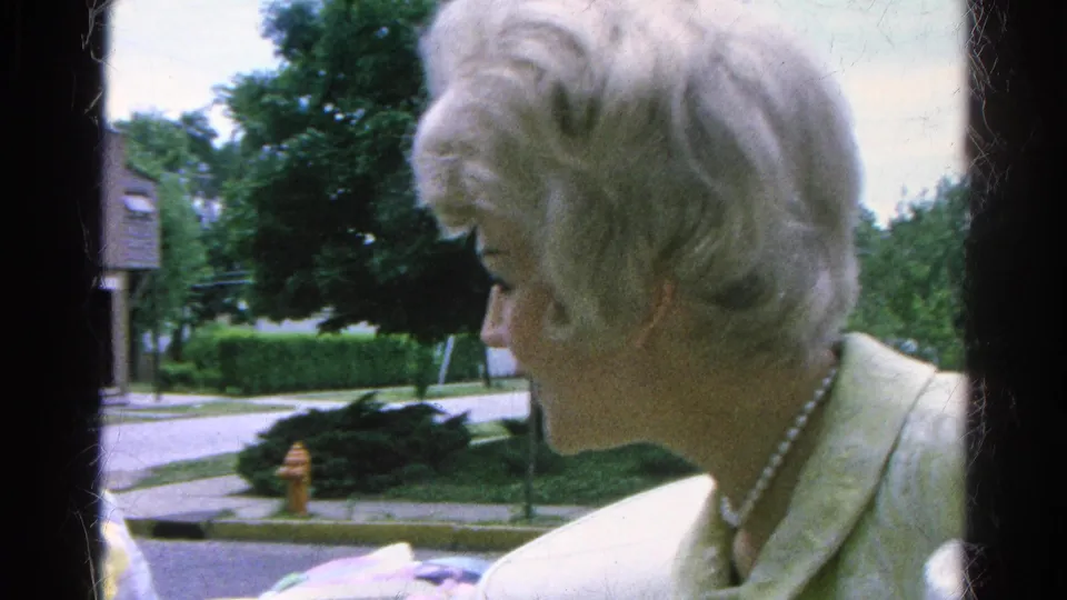 In a 1966 Danville residential setting, a blonde woman in a white jacket and pearls appears in profile near the street.