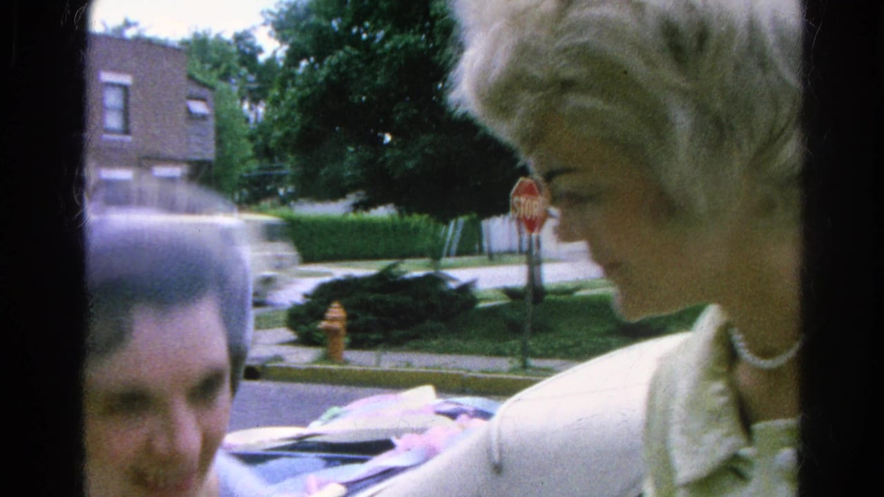 Woman in light coat and pearl necklace near a sidewalk, with a partially visible man and suburban street behind her.