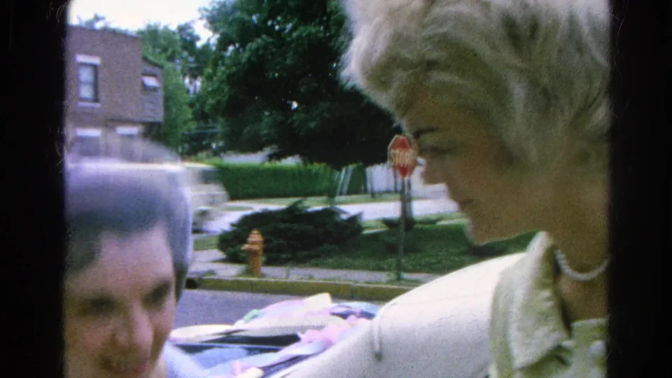 In 1966 Danville, Illinois home-movie footage, a woman in a light coat and pearl necklace stands near a sidewalk beside a partly visible man.