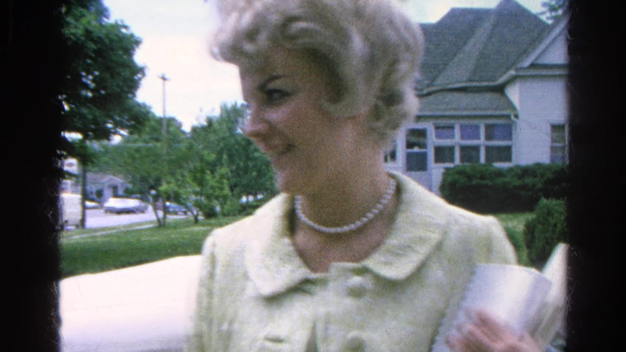Smiling woman in a light coat holds papers or a booklet on a sidewalk with houses and trees behind her.