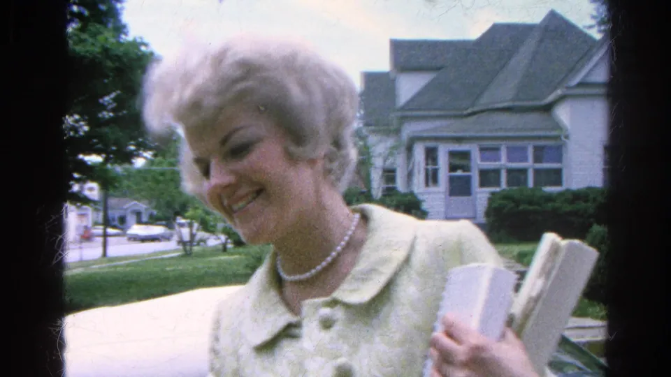 The woman smiles toward the camera in Danville, Illinois, holding papers beside a sidewalk with a house, lawn, and trees behind her.