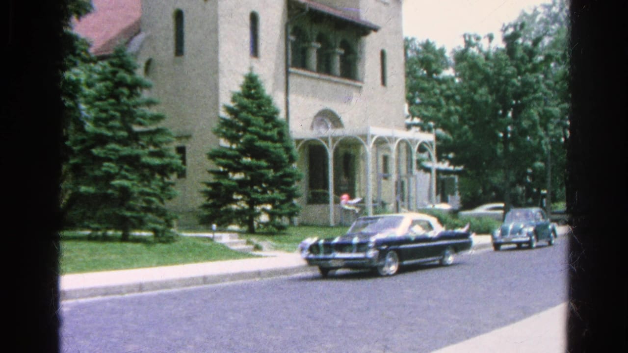 Classic cars pass a church exterior on a sunny neighborhood street.